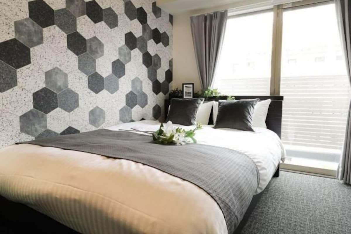 Una habitación de hotel moderna con un muro de estampado geométrico, una cama grande con ropa de cama gris y vistas a la ciudad a través de la ventana.
