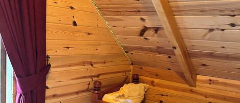 A wooden cabin room with a sloped ceiling, a wooden bed, and a window with curtains.