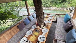 An outdoor dining area with a long wooden table set for a meal, surrounded by cushioned seating under a tree. - Nehir Bungalows (Camlihemsin)
