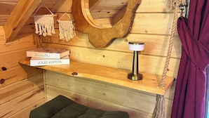 A cozy cabin interior with a wooden ceiling, a round mirror, a small shelf with books, a green cushioned bench, and a rope decoration. - Nehir Bungalows (Camlihemsin)