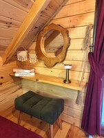 A cozy cabin interior with a wooden ceiling, a round mirror, a small shelf with books, a green cushioned bench, and a rope decoration.