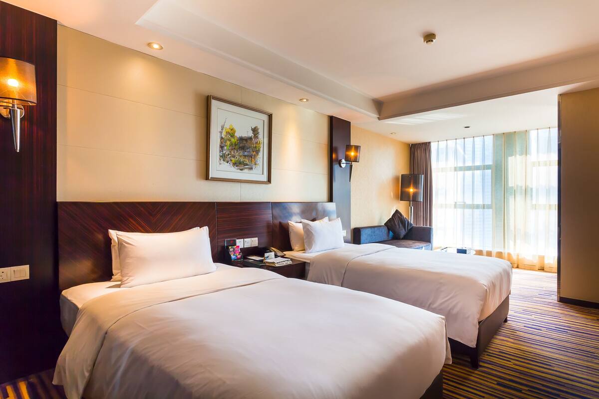 A hotel room with two beds, a nightstand, a lamp, a painting, and a large window.