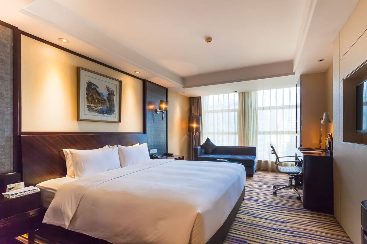 A hotel room with a large bed, a desk with a chair, a sofa, and a large window with curtains.