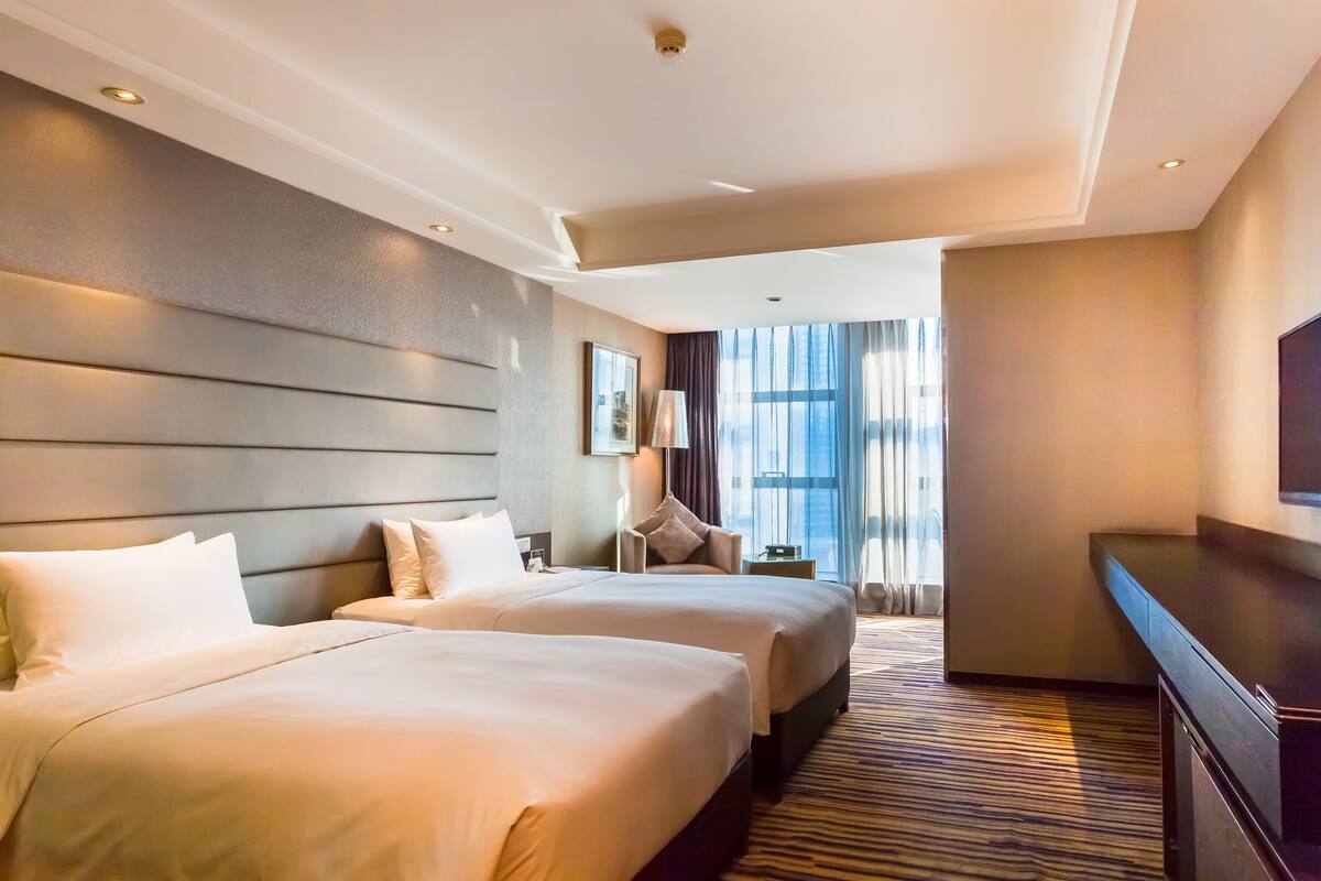 A hotel room with two beds, a desk, a chair, and a large window with curtains.