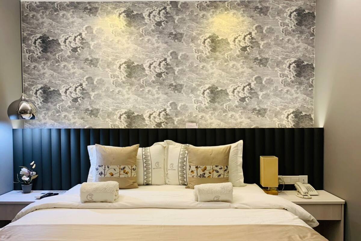 A hotel room with a large bed, a headboard with a cloud pattern, a bedside table with a lamp, and a nightstand with a phone.