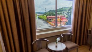 A room with a large window overlooking a traditional building with a red roof. There are two chairs and a small table with a flower vase. - Red City Hotel (Lawas)