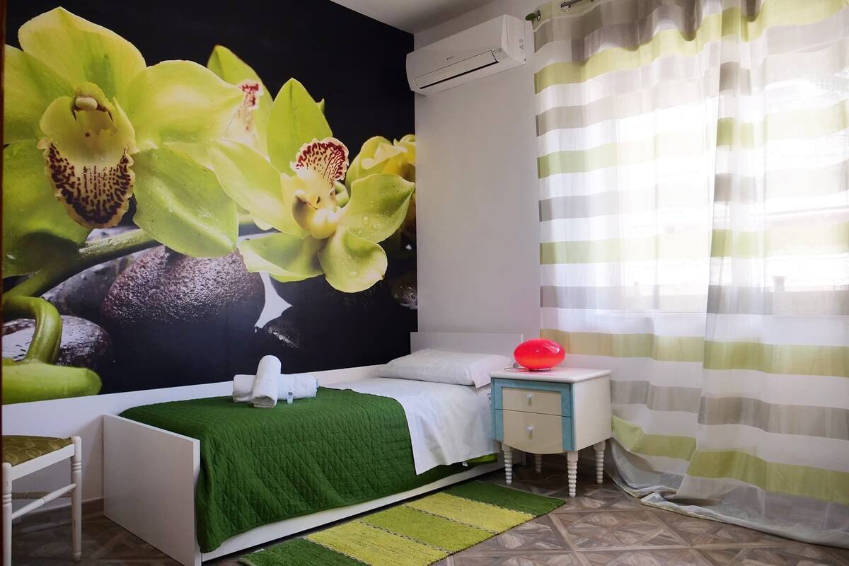 A bedroom with a bed, a bedside table, a nightstand, a green rug, and a wall mural of orchids.