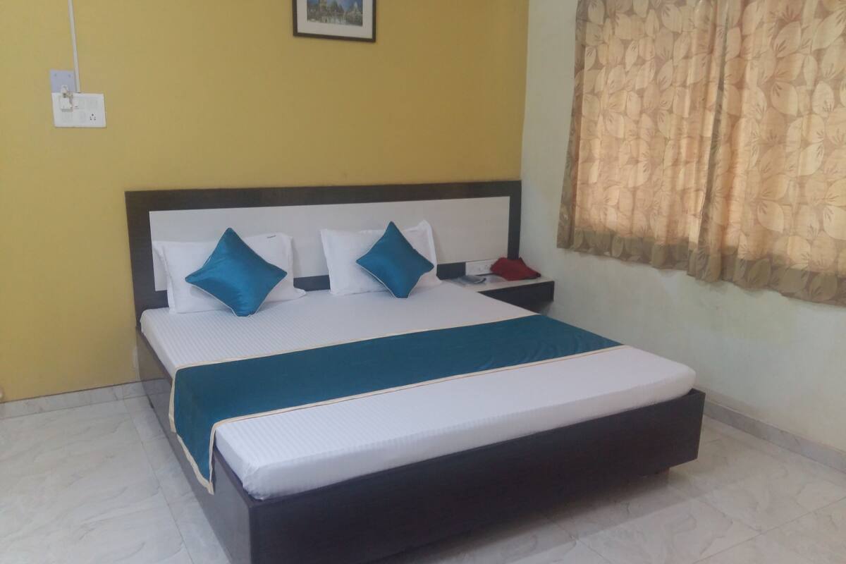 A bed with a white mattress and blue bedding, a bedside table with a red object, and a window with a floral curtain.