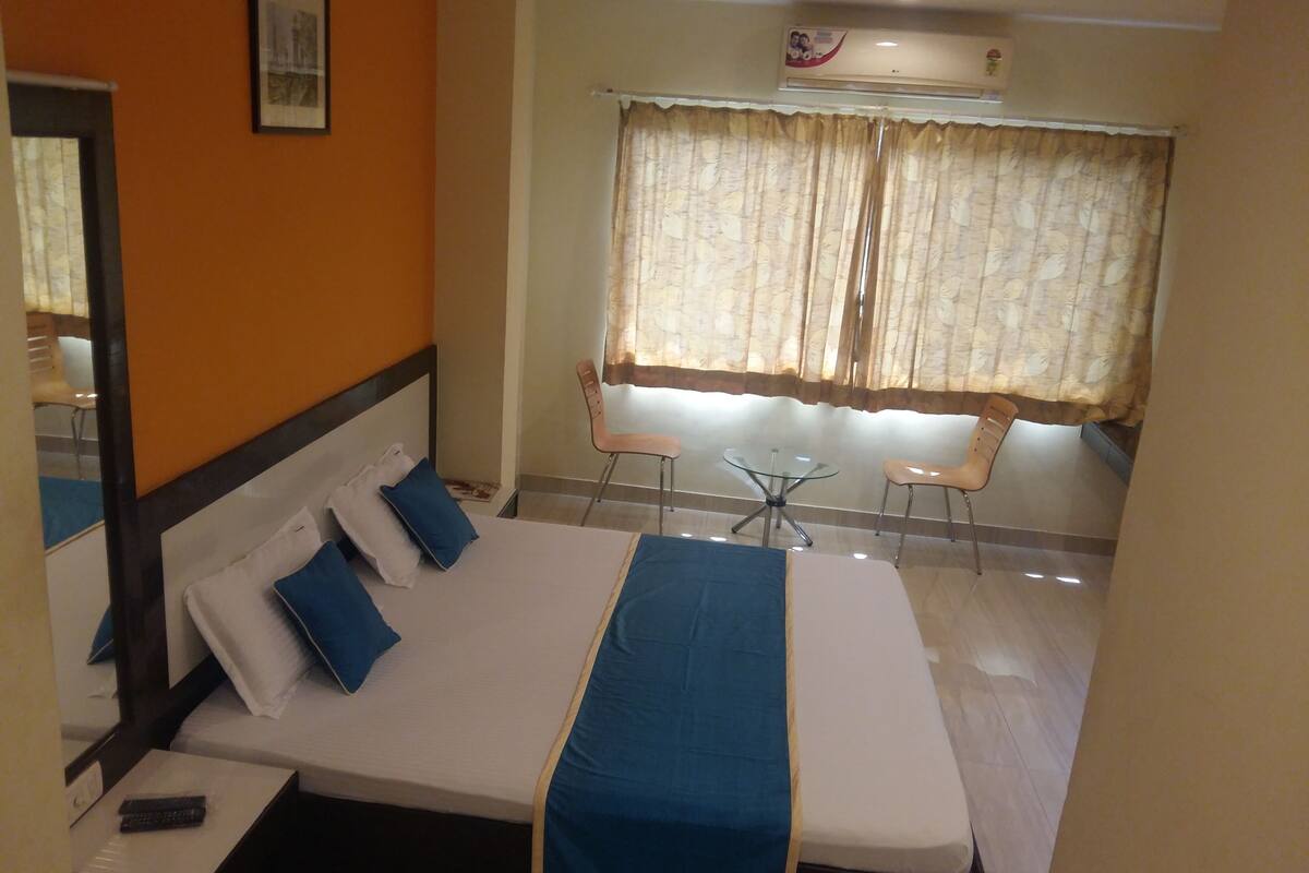A hotel room with a bed, a small table with chairs, a window with curtains, and an air conditioning unit on the wall.
