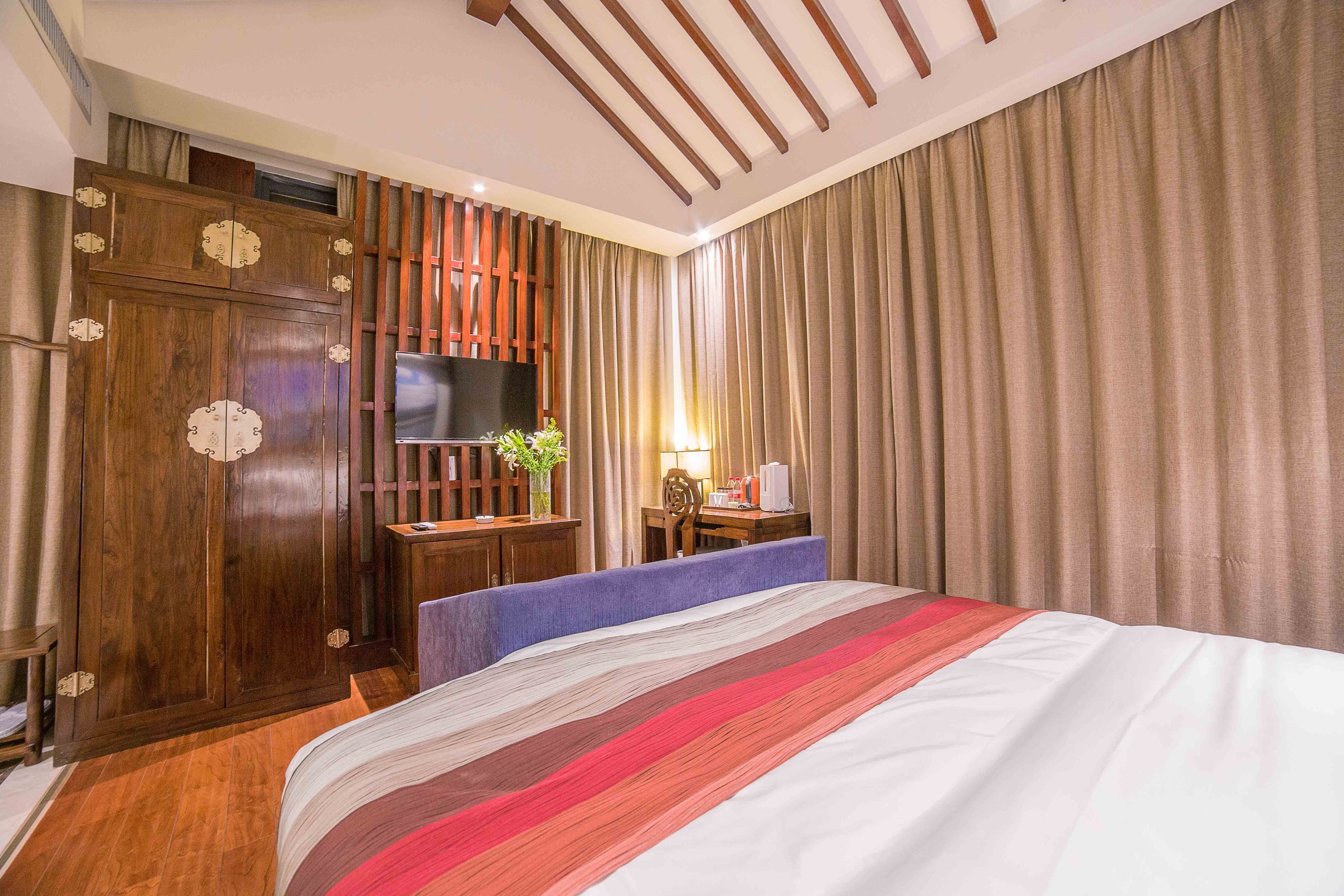 A hotel room with a large bed, wooden wardrobe, a desk with a lamp, and a television.