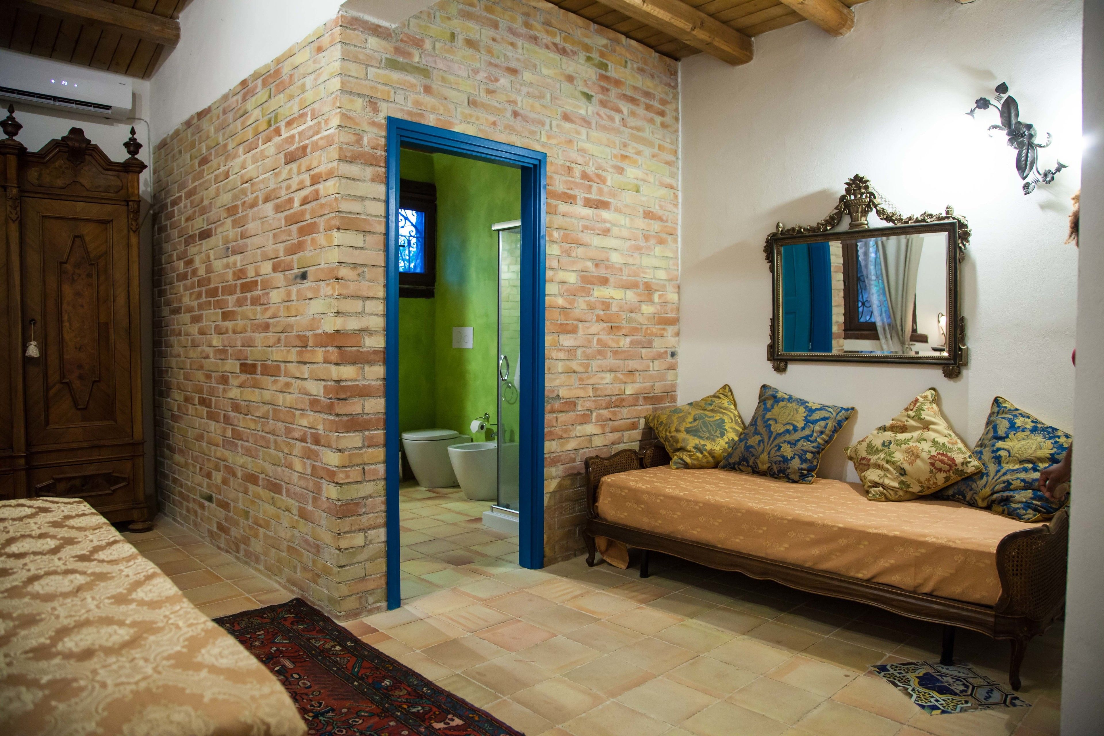 Una habitación con pared de ladrillo, armario de madera, sofá con cojines estampados, espejo y suelo de baldosas.