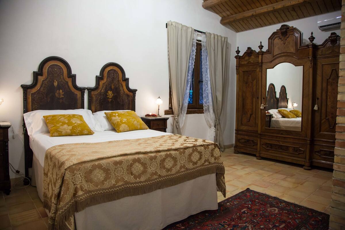 Un dormitorio con una cama grande, cabecera de madera, una mesita de noche, un aparador y una ventana con cortinas.