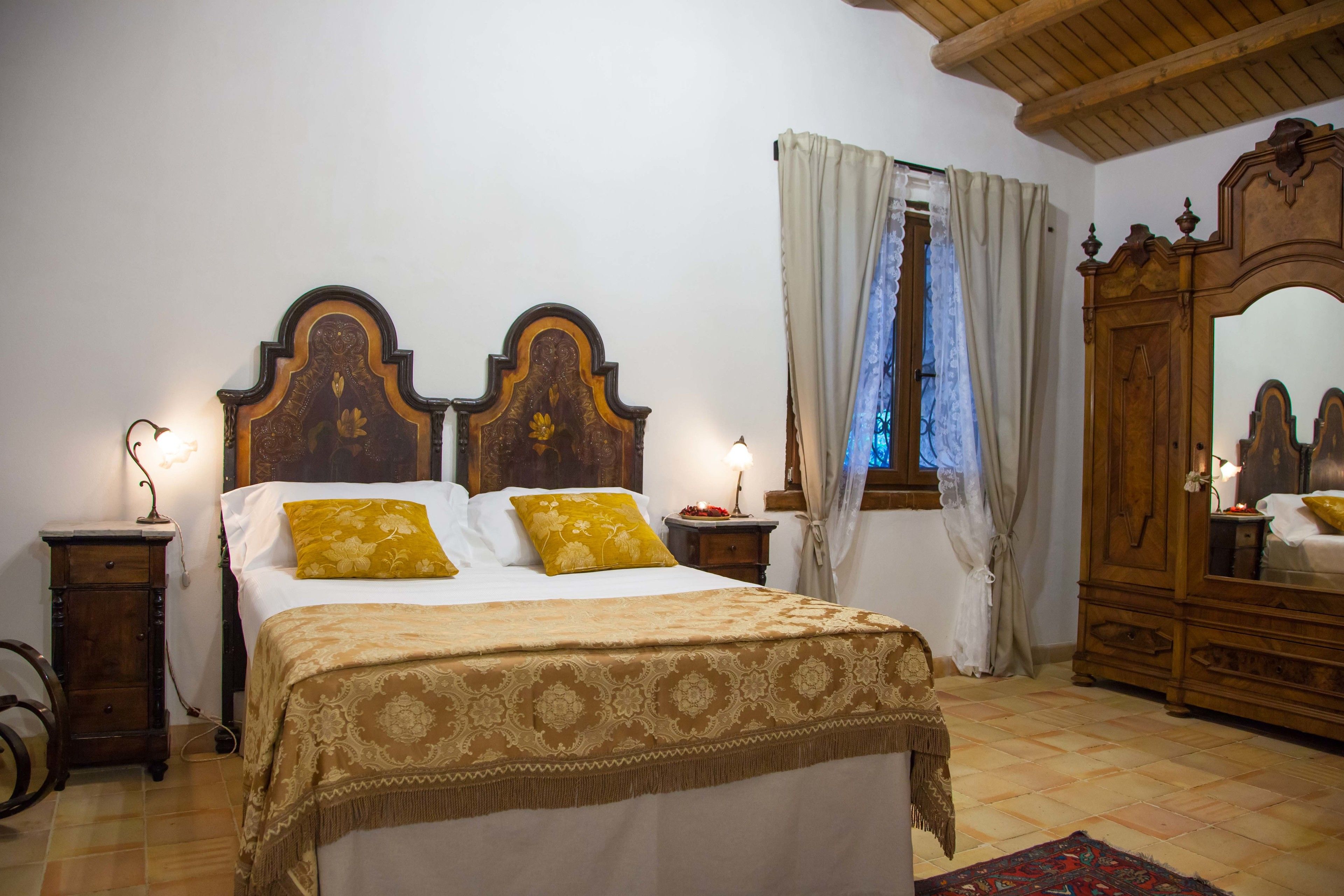 A bedroom with a large bed, wooden headboard, two bedside tables with lamps, a wardrobe, and a window with curtains.