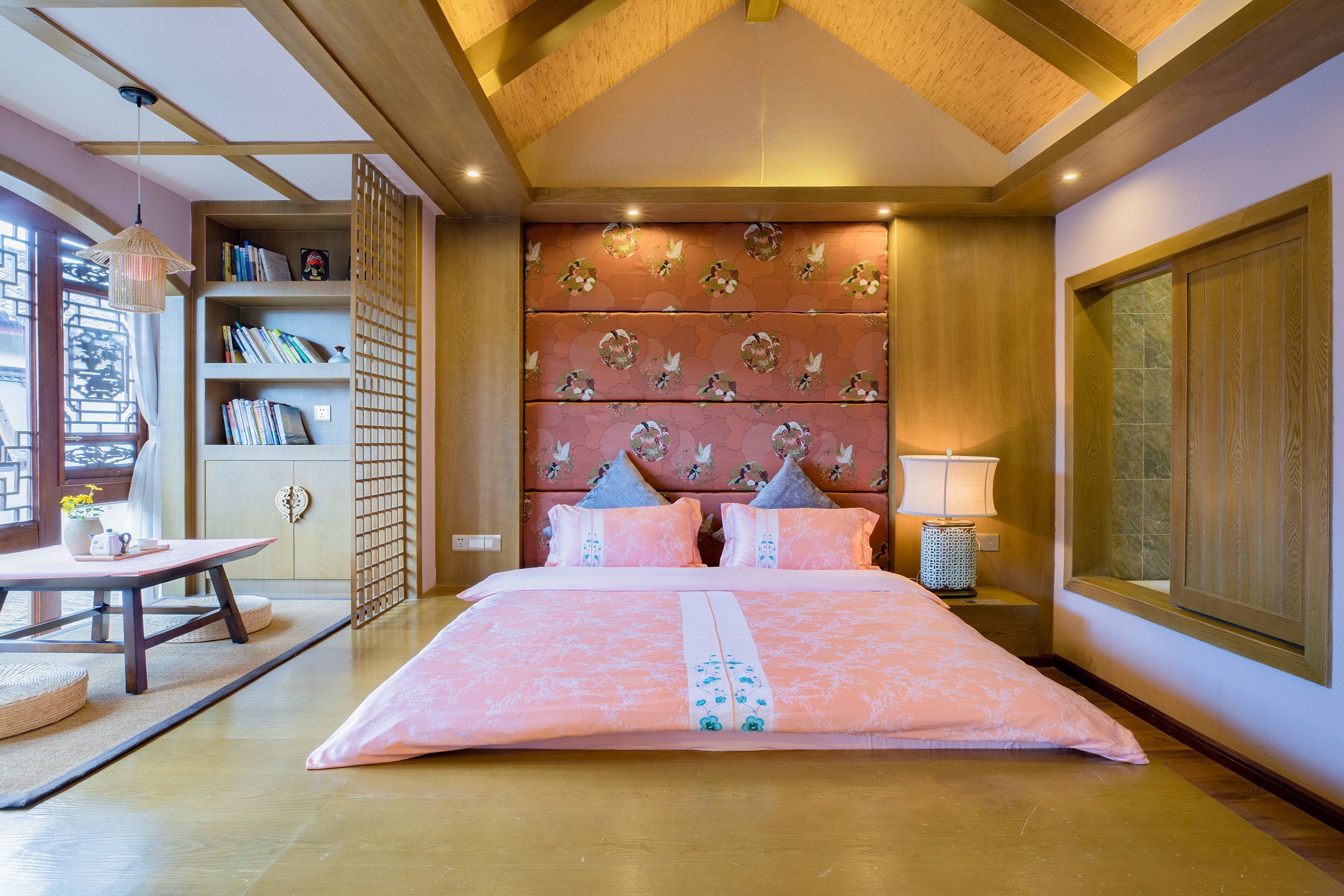 A bedroom with a large bed, a wooden headboard with floral patterns, a bedside table with a lamp, and a bookshelf.