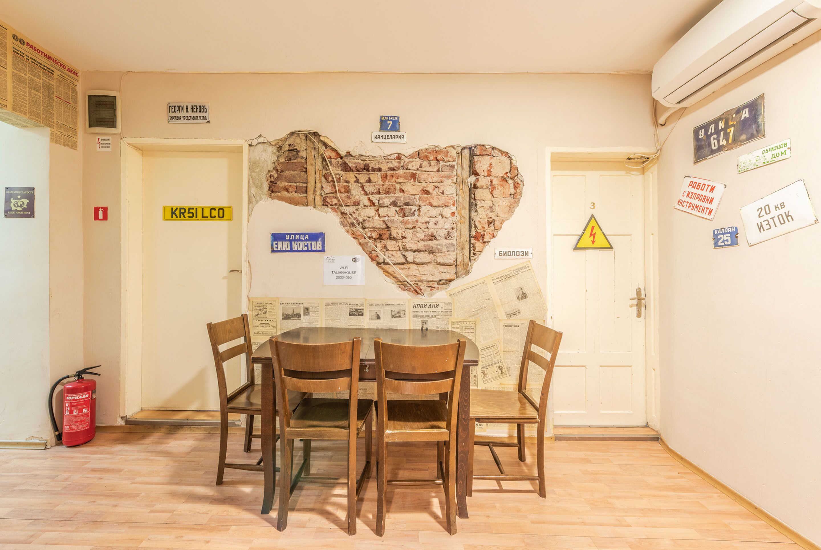 Een eethoek met een muur van bakstenen in de vorm van een hart, houten stoelen en een tafel. Er hangen borden aan de muur, een brandblusser en een deur met een nummer.