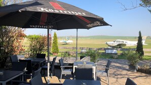 An outdoor seating area with tables and chairs, an umbrella, and a view of an airstrip with planes. - Pension Flying Ranch (Koelleda)
