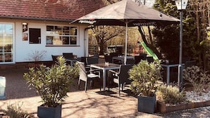 An outdoor seating area with tables and chairs under a large umbrella, surrounded by potted plants. - Pension Flying Ranch (Koelleda)