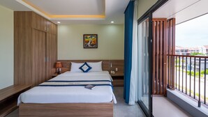 A hotel room with a large bed, a wooden wardrobe, a bedside table with a lamp, a painting on the wall, and a balcony with a view of buildings. - Sofia Hotel (Phu Quoc)
