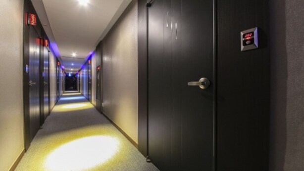 Een hotelgang met een donkere deur aan de rechterkant, een verlichte vloer, en een rij deuren aan de linkerkant.