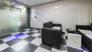 Een hotelkamer met een geruite vloer, twee zwarte banken, een kleine tafel en een glazen scheidingswand.