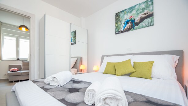 Een hotelkamer met een bed, twee kussens, een nachtkastje, een lamp, een spiegel, een bank en een raam.
