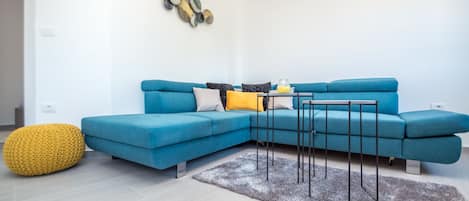 A modern living room with a blue sectional sofa, a yellow pouf, a grey shag rug, and a wall-mounted air conditioner.