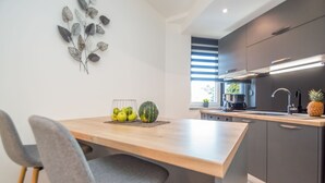 Een moderne keuken met een houten tafel, twee grijze stoelen, een fruitschaal met appels en een watermeloen, en een ananas op het aanrecht.