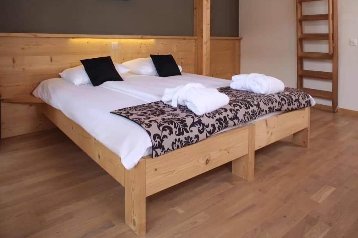 Ein Bett mit Holzrahmen, weiße Bettwäsche und einer gemusterten Decke, ein gerahmtes Bild an der Wand und eine Holzleiter.