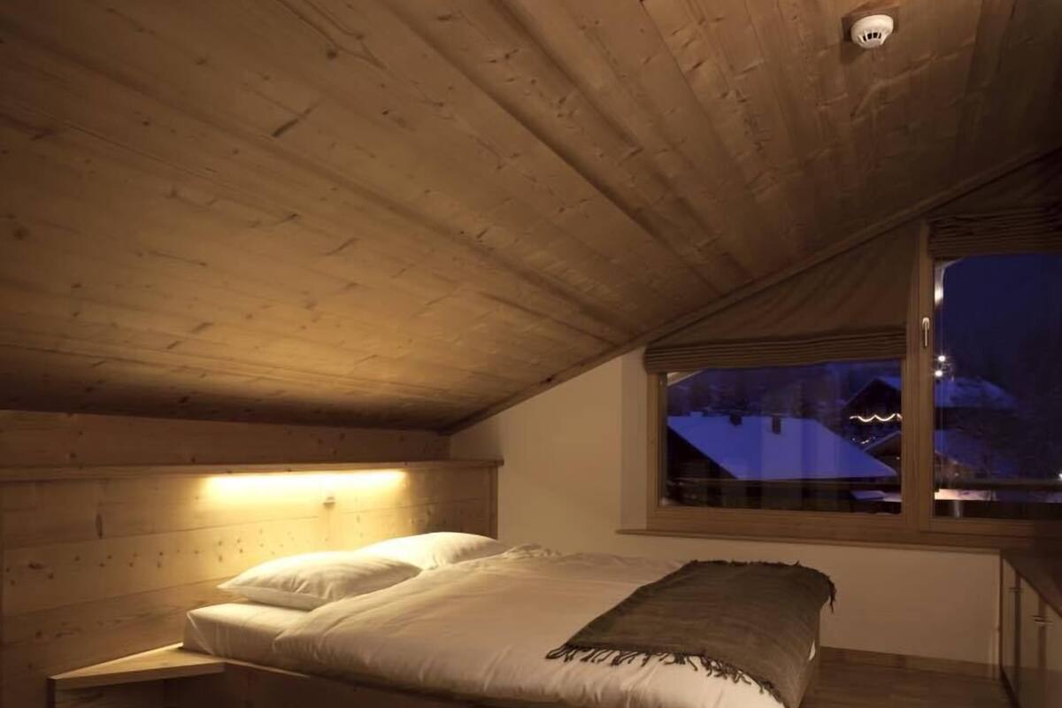 Ein Schlafzimmer mit Holzboden, einem Bett mit weißer Bettwäsche und einem Fenster mit Blick auf eine verschneite Landschaft.
