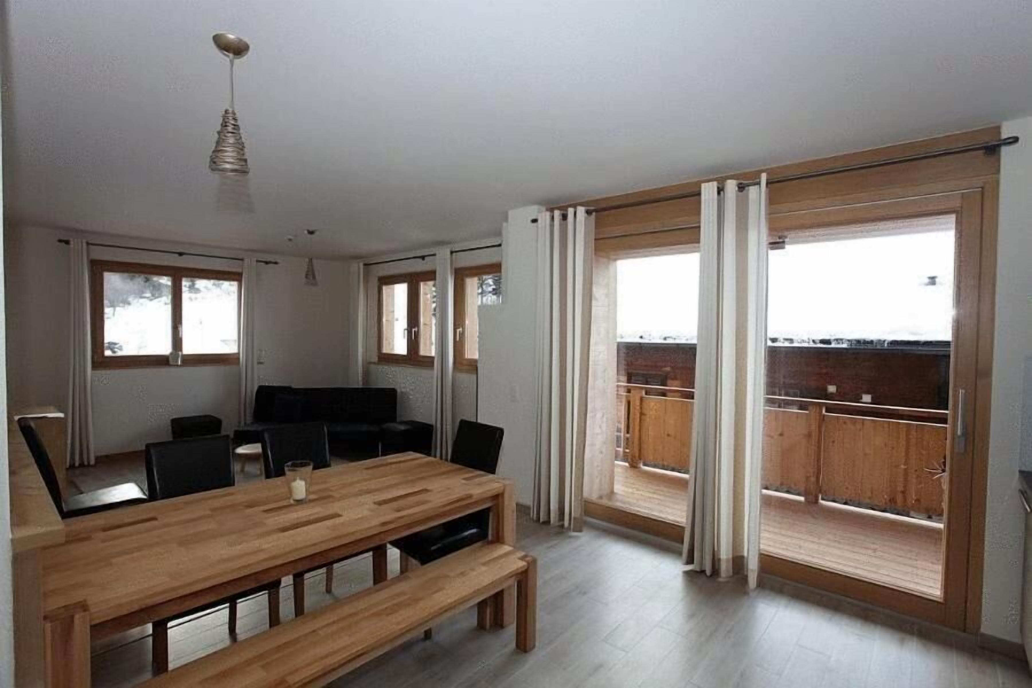 A spacious room with a wooden dining table, chairs, and a bench. There are large windows with curtains, a ceiling light, and a view of a balcony with wooden railings.