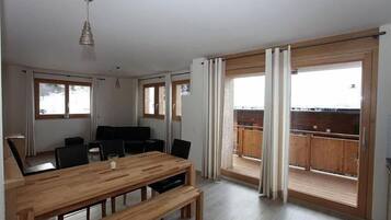 A spacious room with a wooden dining table, chairs, and a bench. There are large windows with curtains, a ceiling light, and a view of a balcony with wooden railings.