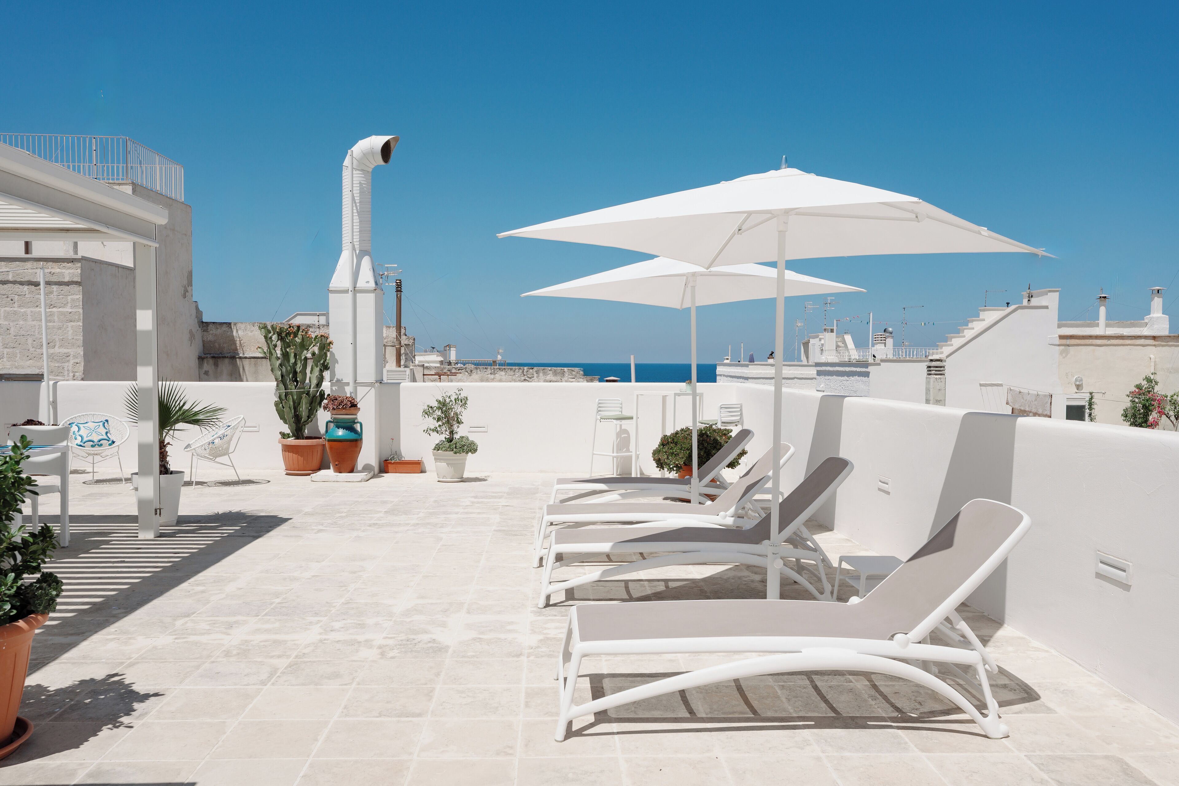 Terraza en la azotea con sillones blancos, sombrillas, plantas en macetas y una vista despejada al mar.