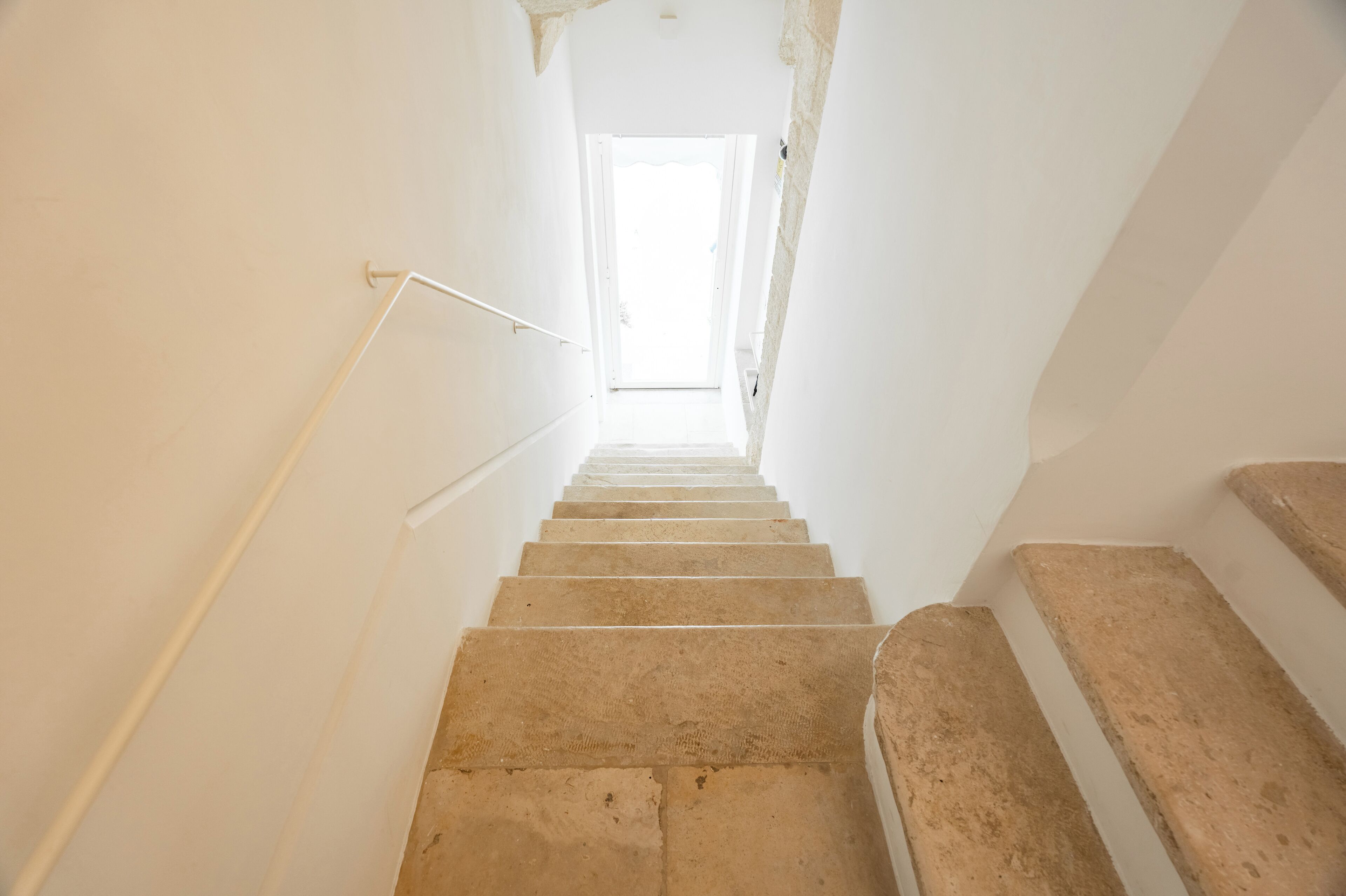 Una escalera que lleva a una habitación luminosa con paredes blancas y una puerta visible en la parte superior.