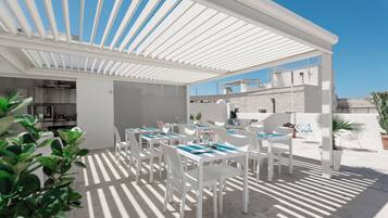 Un espace extérieur abrité avec des tables et des chaises blanches, une cuisine à l’arrière-plan et un ciel bleu dégagé.