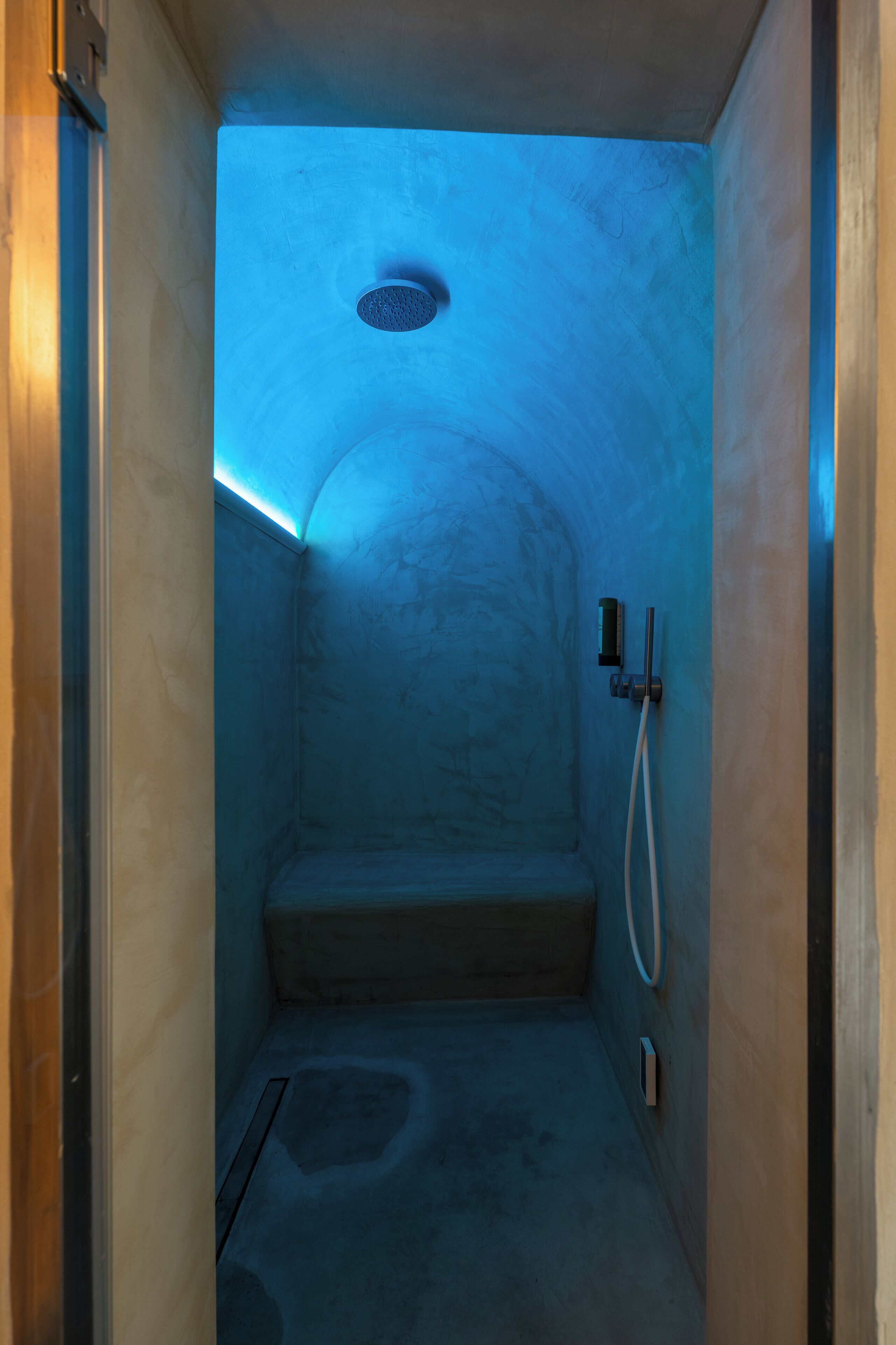A small, enclosed bathroom with a blue light, a showerhead, and a step leading to a shower area.