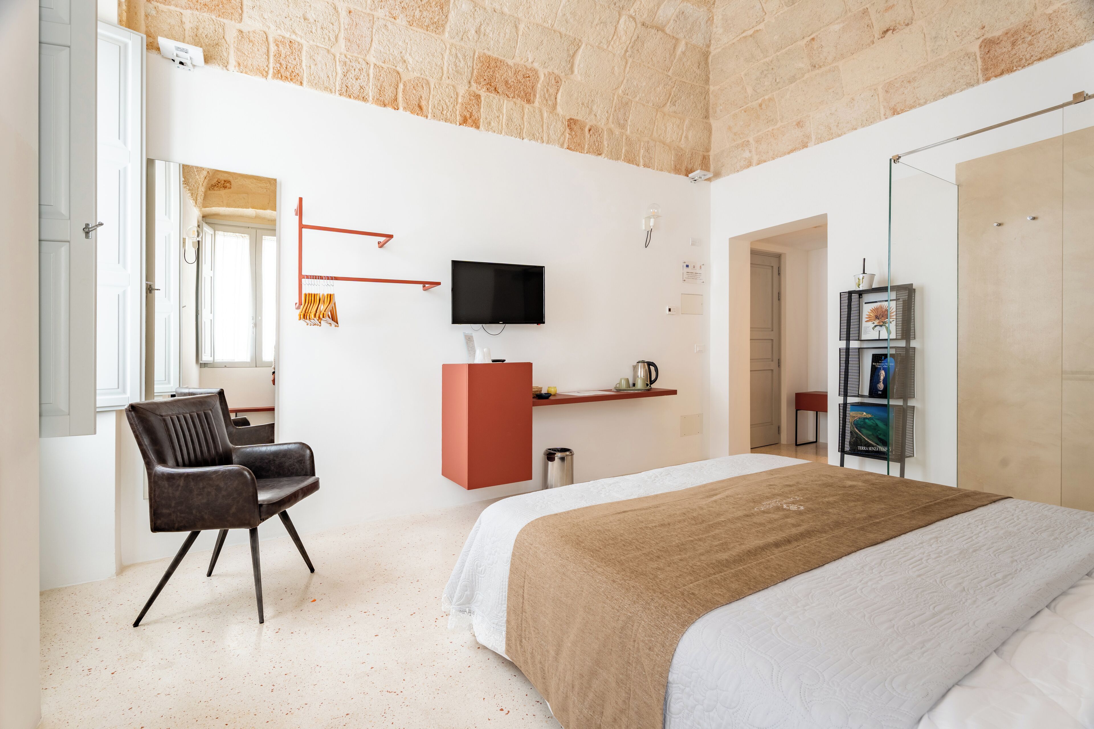 A modern hotel room with a large bed, a seating area with a chair, a TV mounted on the wall, and a stone wall.