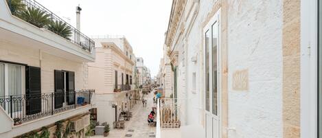 Una calle estrecha flanqueada por edificios, con una persona caminando y zonas de estar al aire libre.