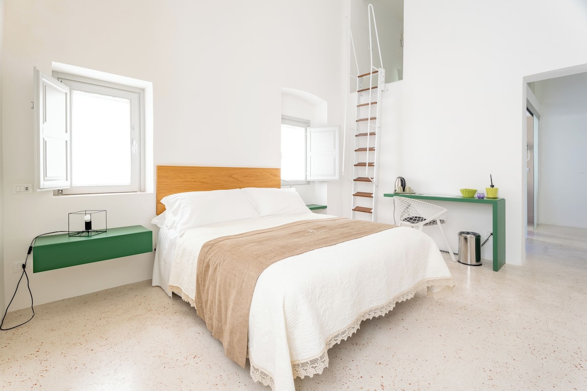A bedroom with a bed, a green nightstand, a desk with a chair, a ladder, and a window.