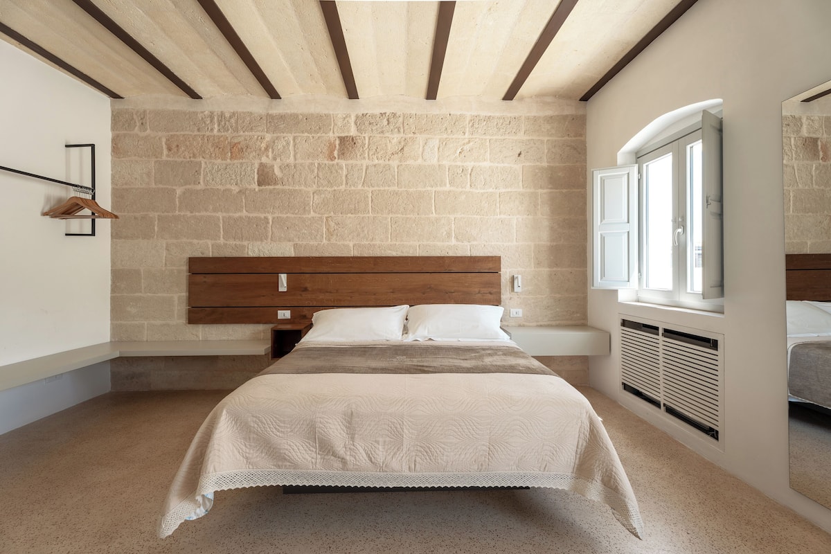 A bedroom with a stone wall, wooden headboard, and a window with blinds.