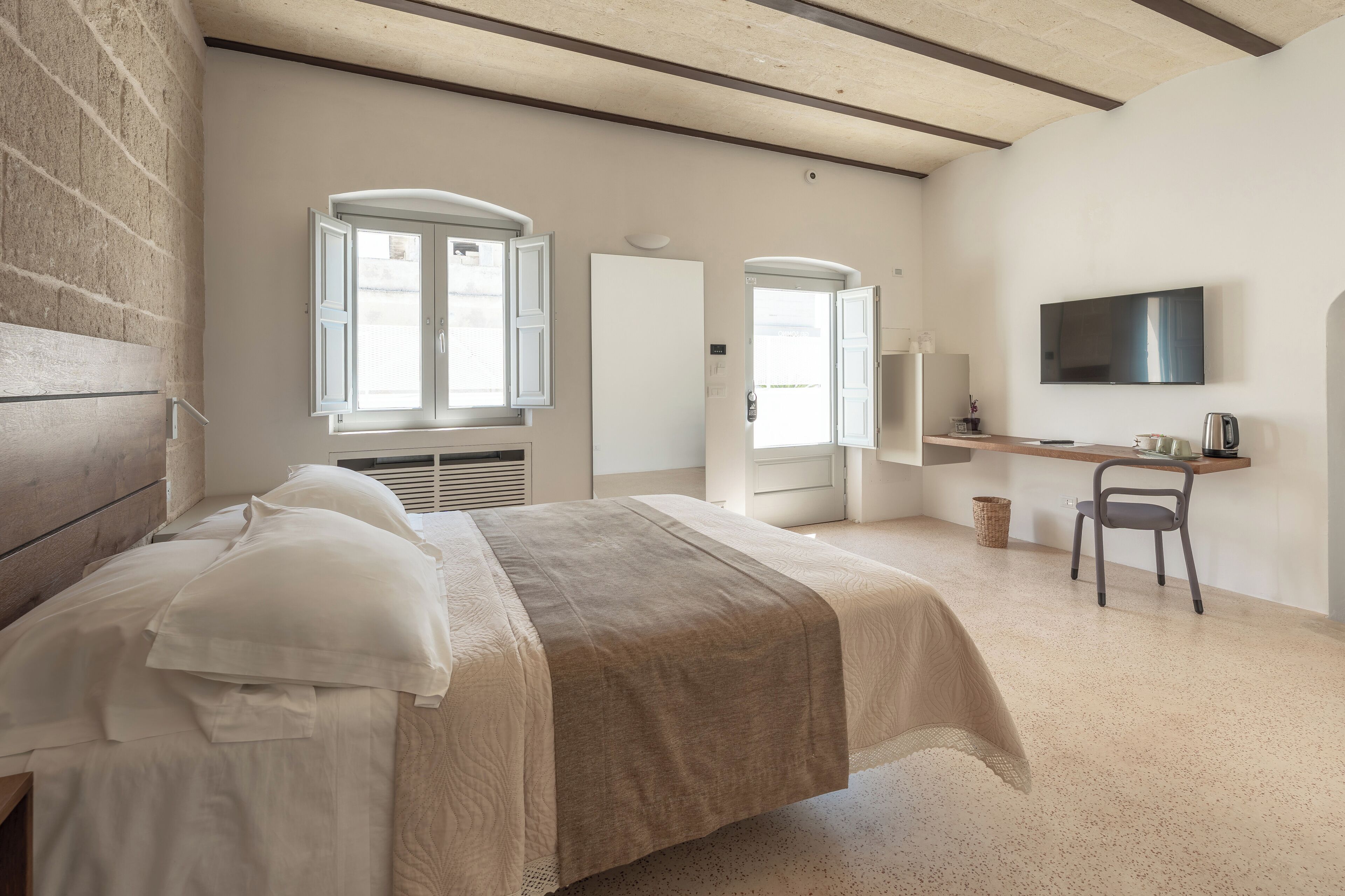 Ein Schlafzimmer mit einem Bett, einem Schreibtisch, einem Sessel, einem Fernseher und einem Fenster mit geöffneten Lamellen.