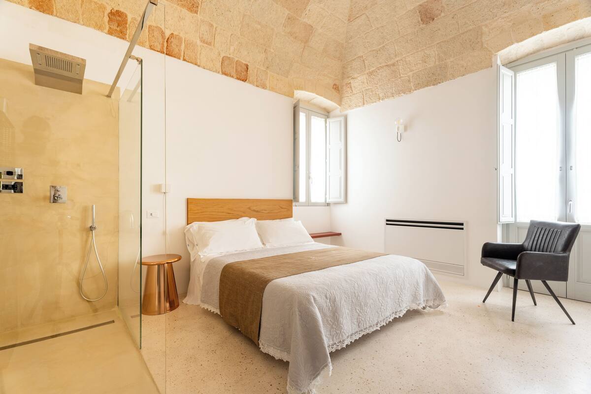 A bedroom with a bed, a chair, a shower, and a window.
