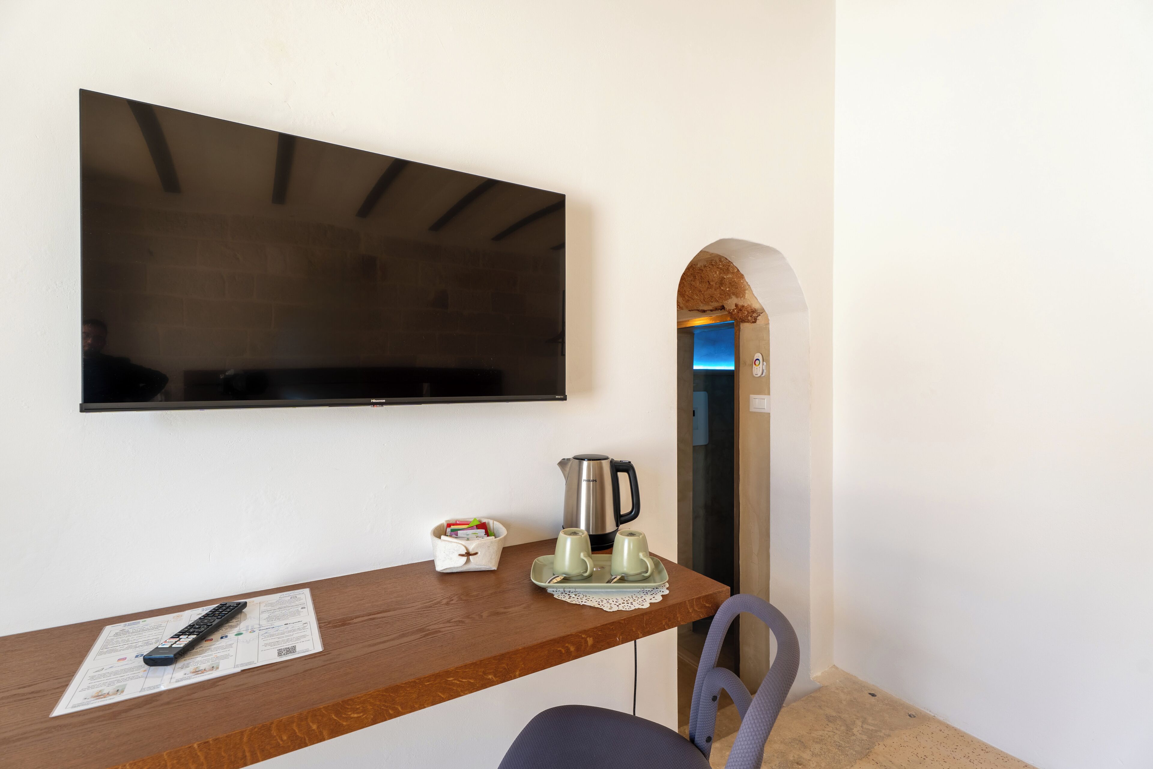 Een kamer met een wandtelevisie, een houten tafel met een afstandsbediening, een waterkoker en een dienblad met twee kopjes.