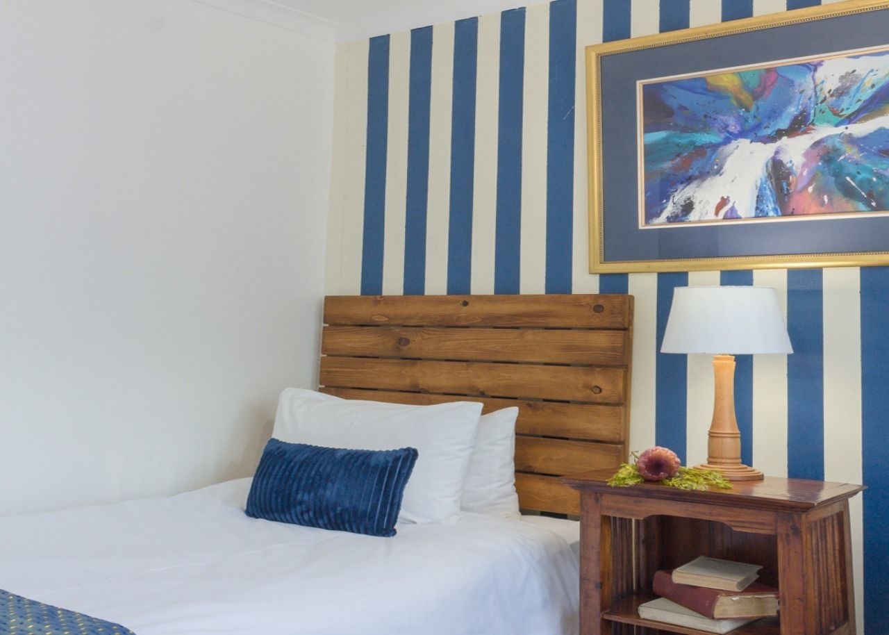 A hotel room with a wooden headboard bed, white bedding, a blue pillow, a bedside table with a lamp, and a framed abstract painting on the wall.