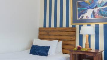 Habitación de hotel con cama de cabecera de madera, ropa de cama blanca, almohada azul, mesita de noche con lámpara y un cuadro abstracto enmarcado en la pared.