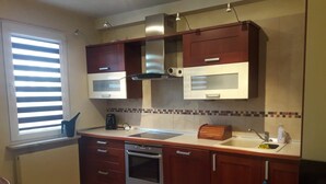 A compact kitchen with wooden cabinets, a built-in oven, a microwave, and a window with blinds. - Apartament Królewski Sandomierz (Sandomierz)