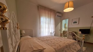 A neatly made bed with a patterned bedspread and pillows, a white radiator, a window with sheer curtains, and a television on a stand. - Catarina (Tollo)