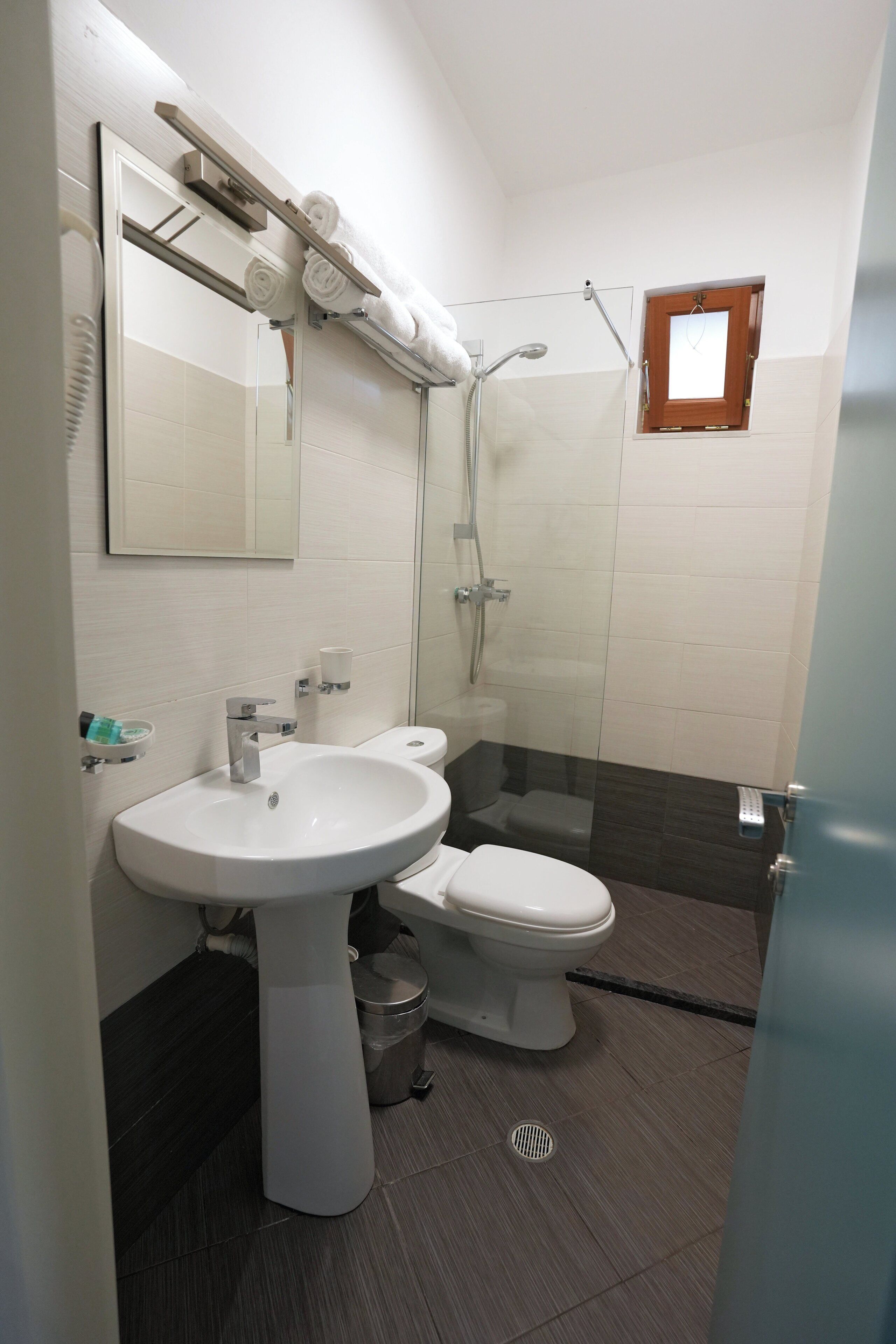 A bathroom with a white sink, toilet, and shower. There is a mirror above the sink and a small window above the shower.