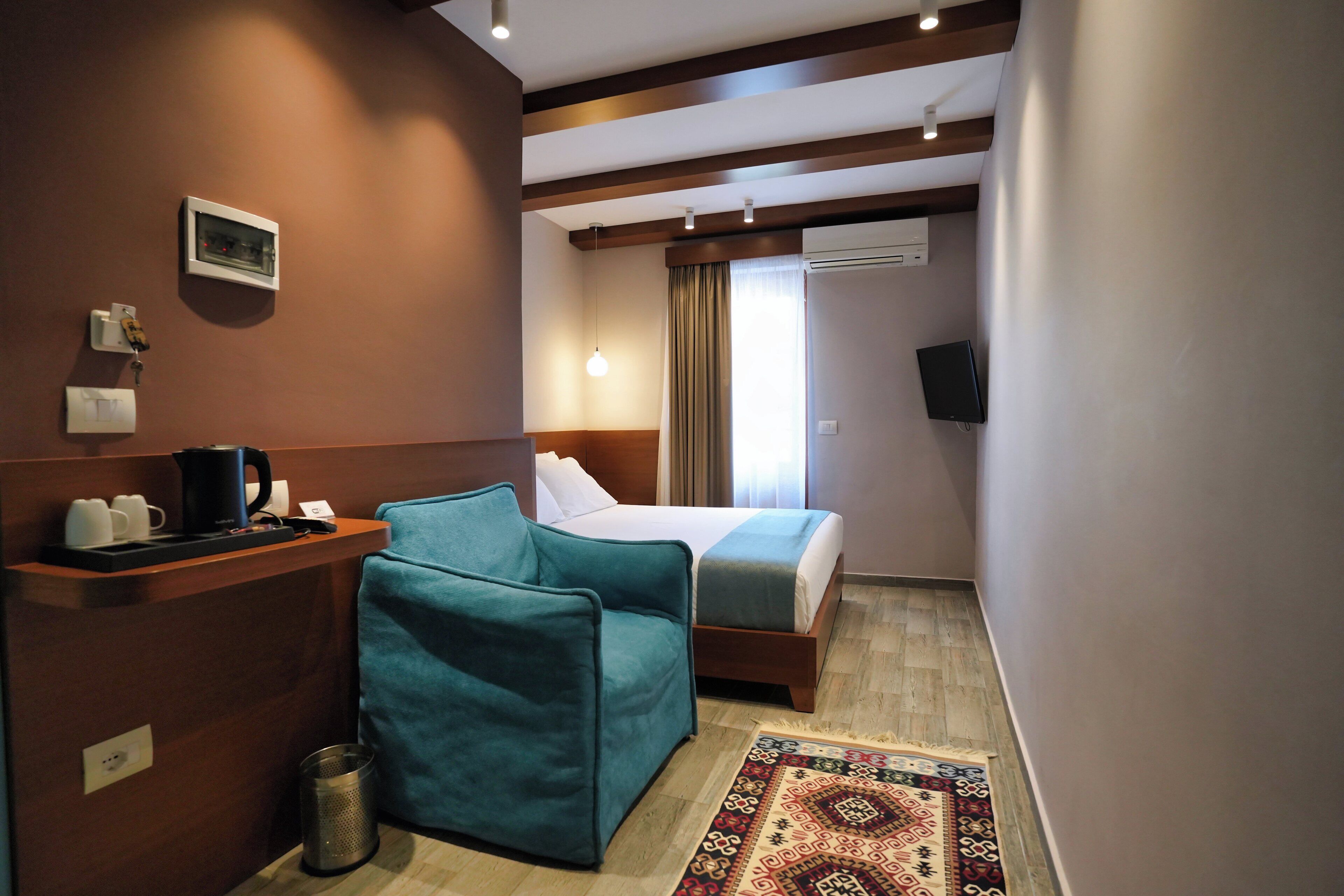 Quarto de hotel com uma cama, uma escrivaninha com cafeteira, um cesto de lixo e um tapete estampado.