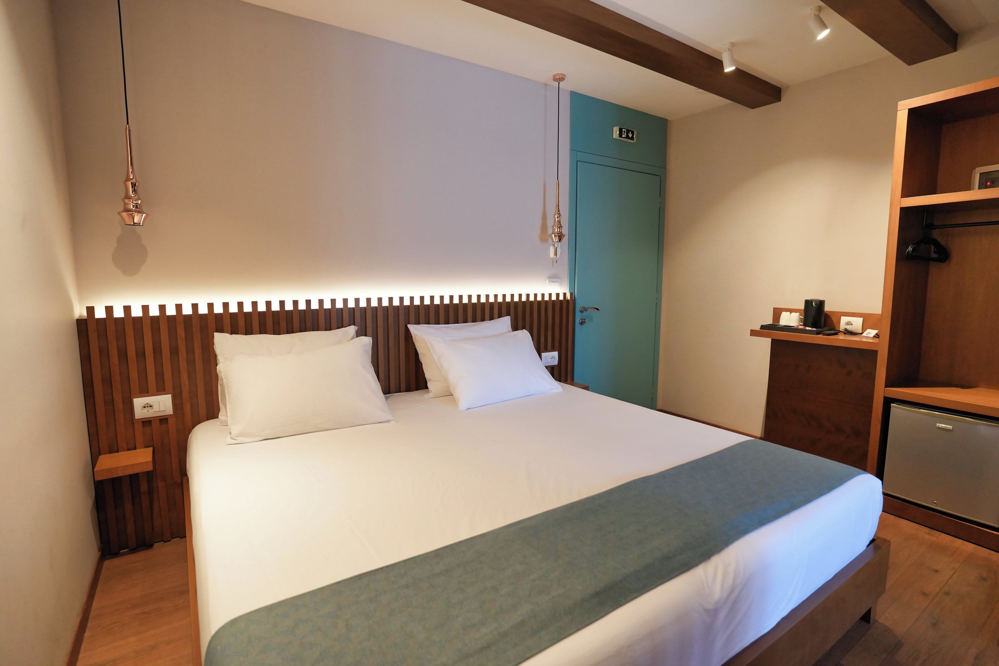 A neatly made bed with a wooden headboard, a teal door, and a small wooden shelf with a kettle.