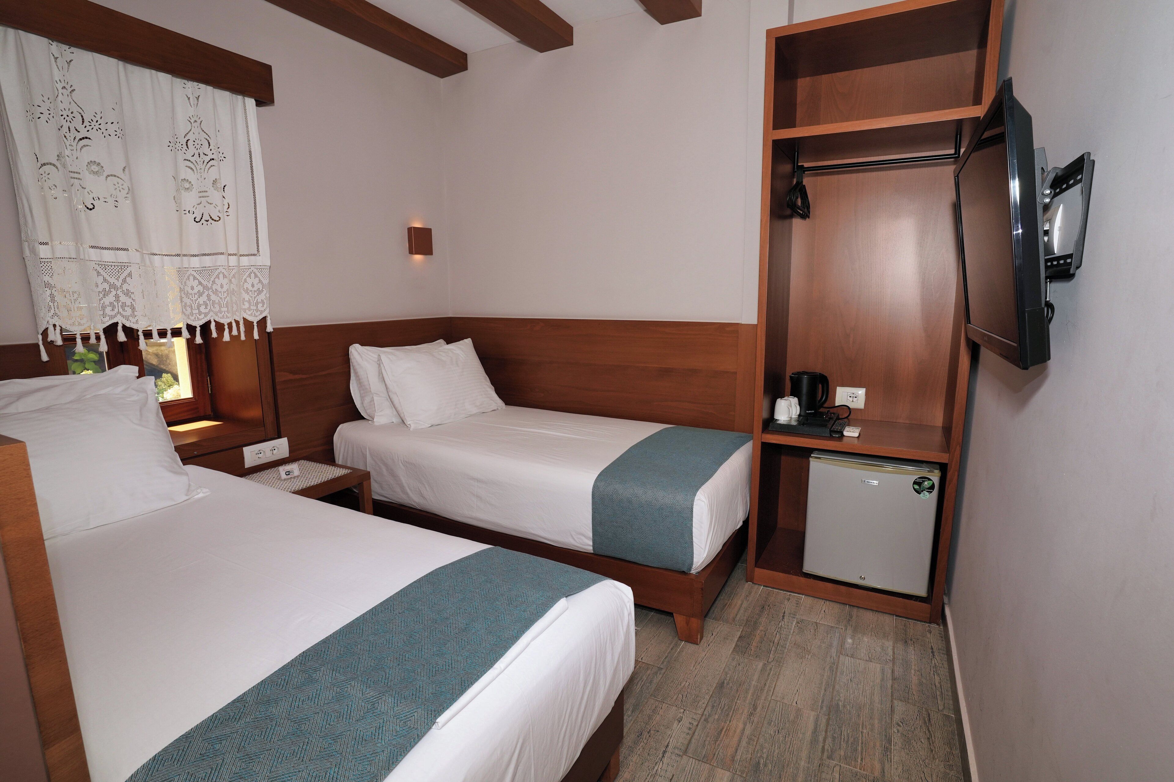 Ein Hotelzimmer mit zwei Betten, einem Holzkopfstück, einem kleinen Kühlschrank, einem an der Wand befestigten Fernseher und einem Fenster mit Vorhang.